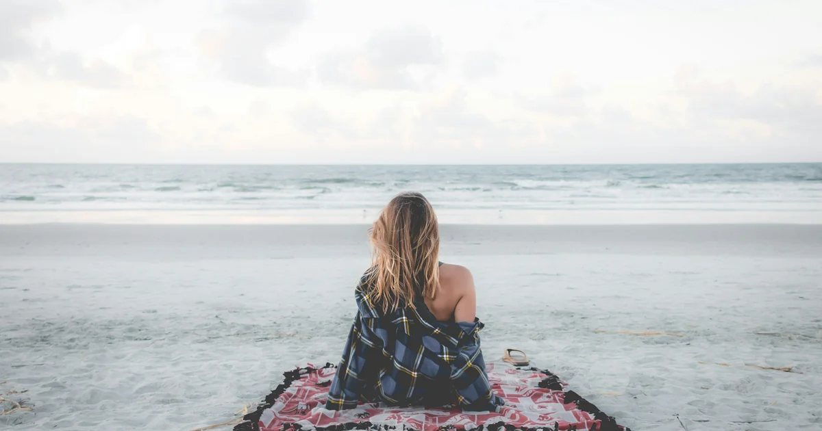Woman wrapped in a blanket sitting quietly on the beach, looking out at the ocean in Huntington Beach near Aloha Recovery.